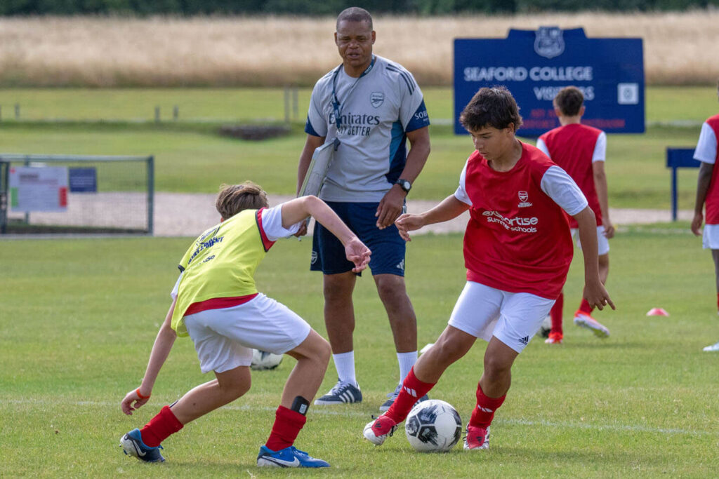 Arsenal camp players training while an Arsenal coach observes the session during a skill-development drill.