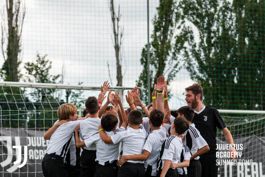 A licensed Juventus soccer camp coach cheering and celebrating with a group of young players during a training session at the 2026 Summer Football Camps in Italy.